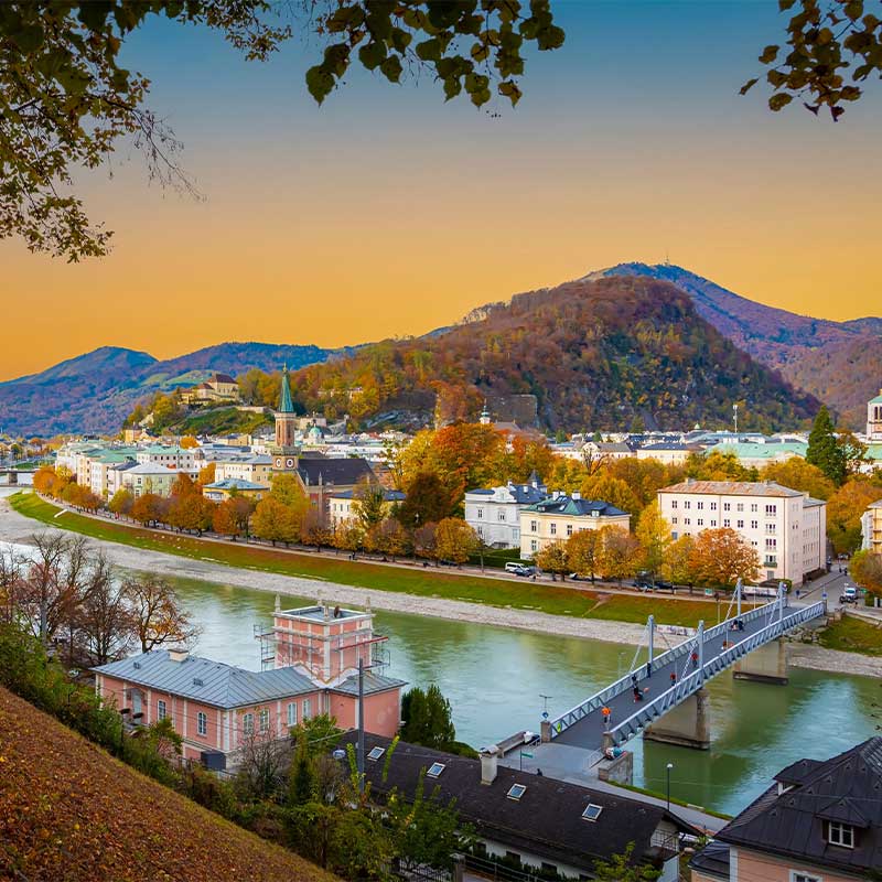 Eine malerische Aussicht auf Salzburg, Österreich, mit dem Fluss Salzach, historischen Gebäuden und Hügeln mit Herbstlaub unter einem leuchtenden Sonnenuntergangshimmel.