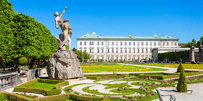 Formal garden with intricate hedges and flowerbeds, featuring a statue and a large historic building in the background under a clear blue sky.