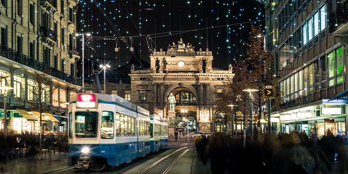 A blue and white tram moves through a busy, illuminated street at night, with a historic building in the background decorated with festive lights.
