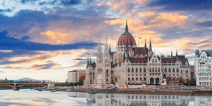 The Hungarian Parliament Building in Budapest is shown against a vibrant sunset sky, with its reflection visible in the Danube River.