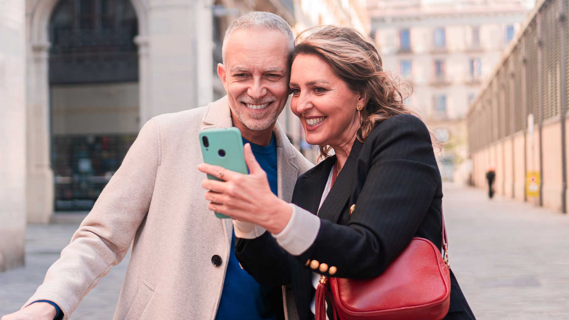 A man and a woman smile while taking a selfie together on a street with buildings in the background.