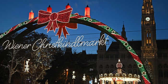 Archway with festive lights and "Wiener Christkindlmarkt" text at an evening Christmas market, with a carousel and illuminated town hall in the background.