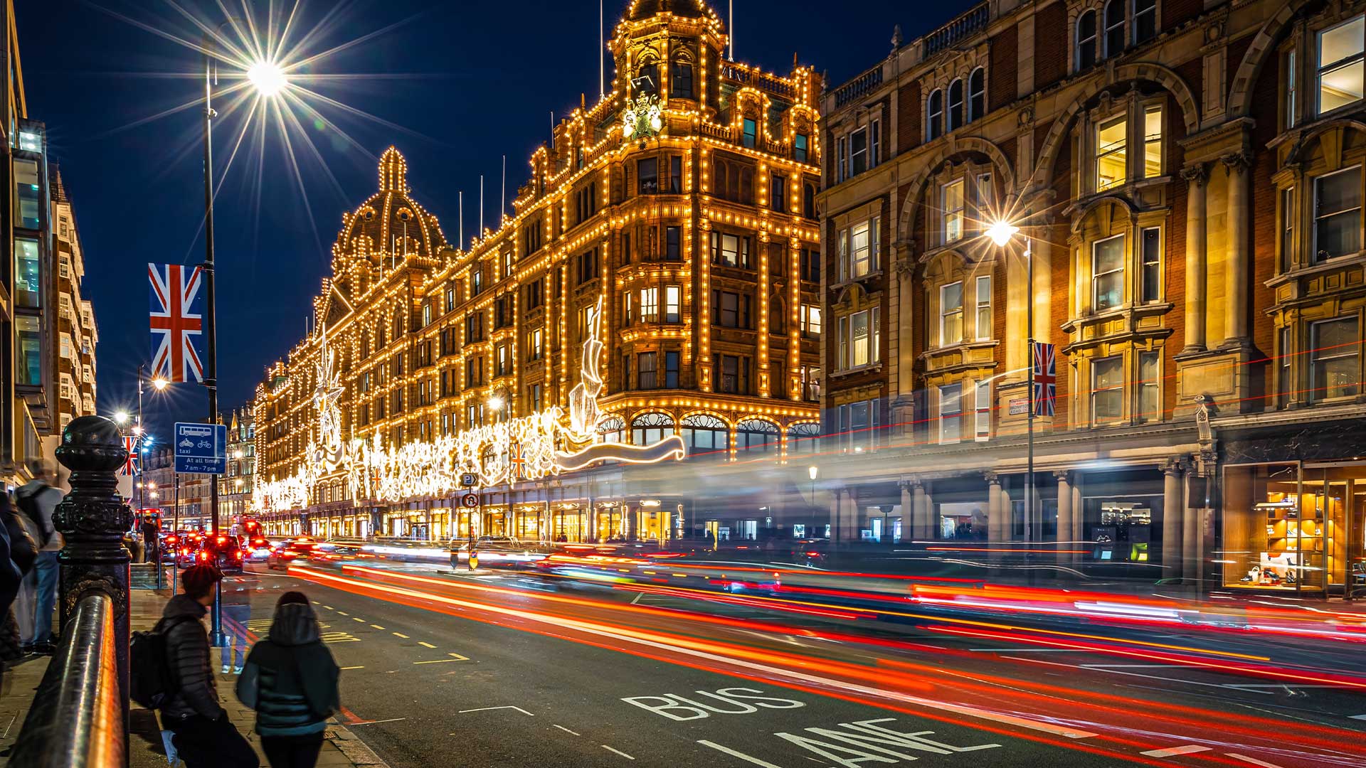 A bustling city street at night, with the facade of a large, illuminated building and the light trails of passing vehicles.