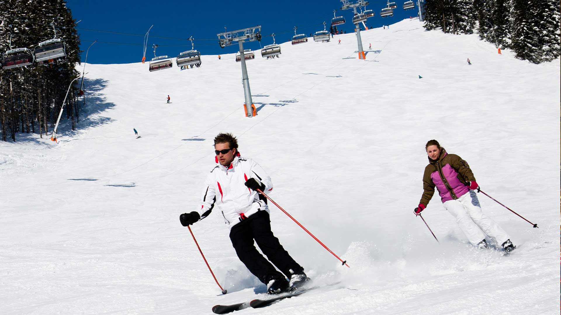 Two skiers in colorful jackets descend a snowy slope under a clear blue sky, with chairlifts and trees visible in the background.