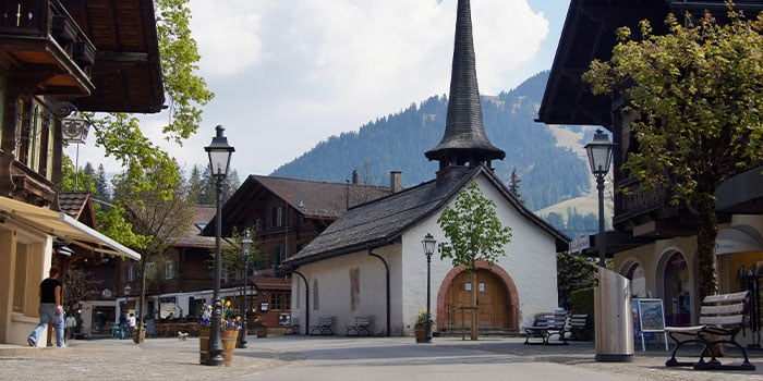 Kleiner Dorfplatz mit weißer Kapelle, spitzem Turm und Holzhäusern. Im Hintergrund sind Berge zu sehen.
