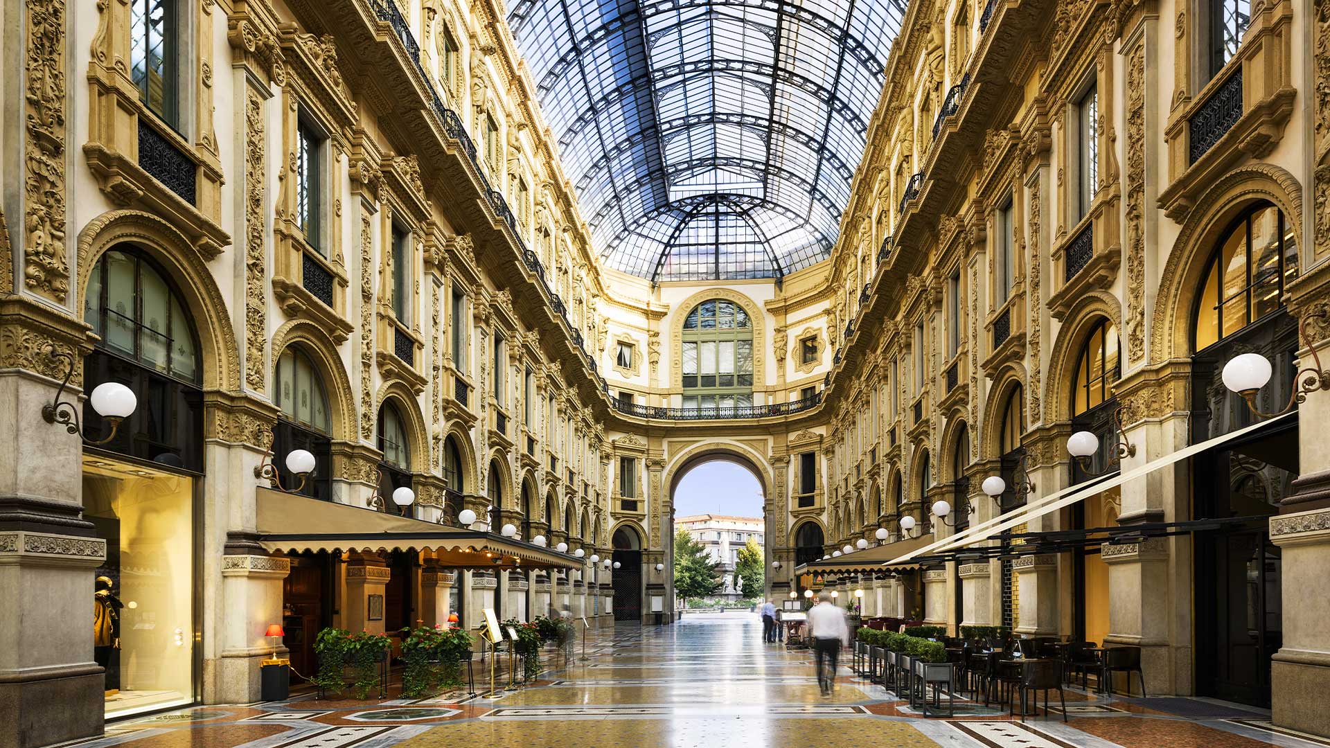 A grand indoor arcade with arched glass ceiling, ornate architecture, and rows of shops and cafes. A few people walk along the patterned floor.