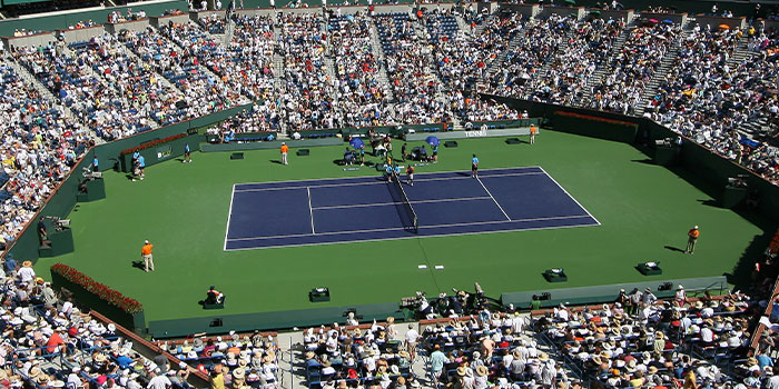 Luftaufnahme eines voll besetzten Tennisstadions während eines Tagesspiels. Der Platz ist grün und die Spielfläche blau. Die Zuschauer drängen sich unter einem klaren Himmel.