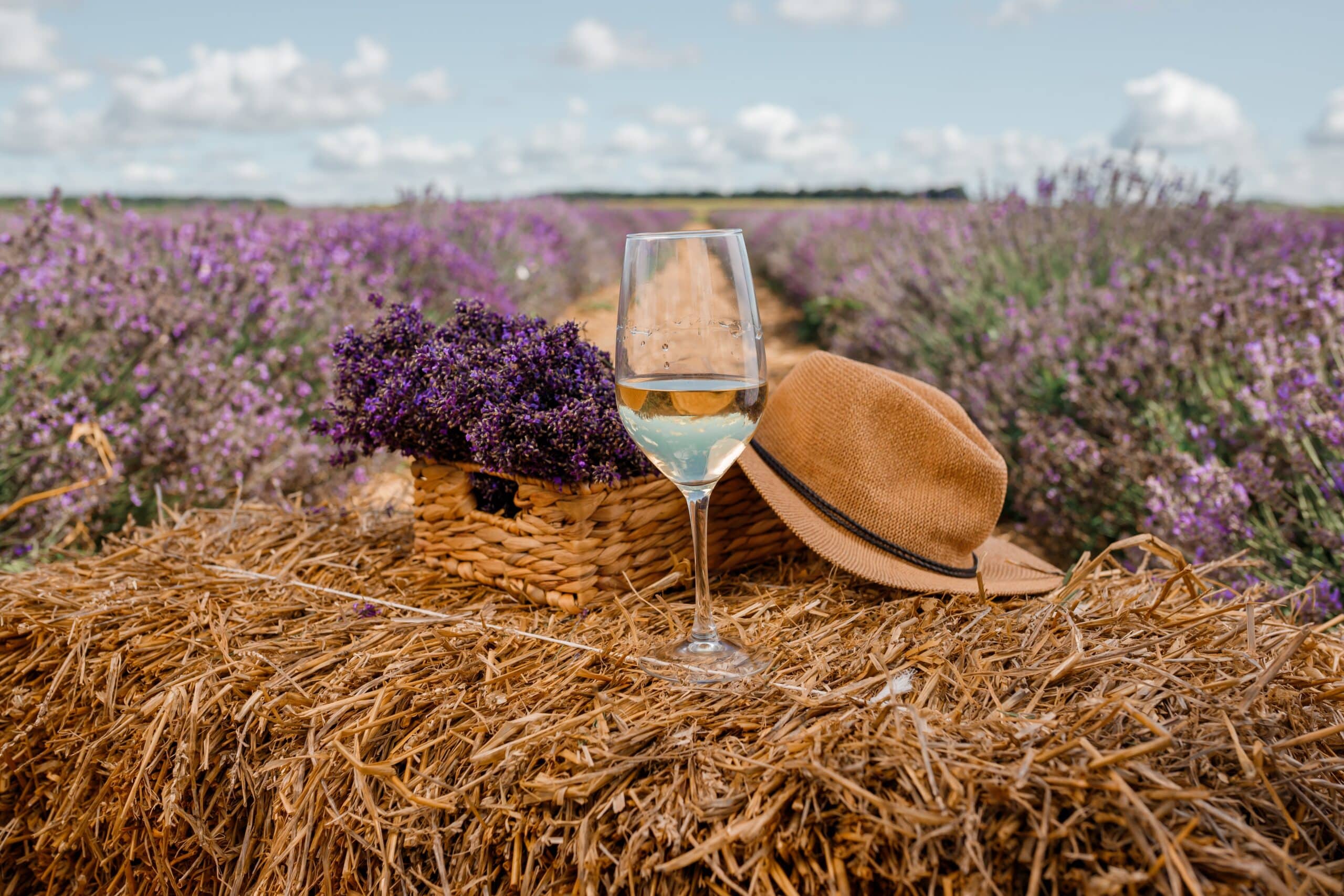 Ein Glas Weißwein, ein geflochtener Korb mit Lavendel und ein Strohhut auf einem Heuballen in einem Lavendelfeld unter einem teilweise bewölkten Himmel.