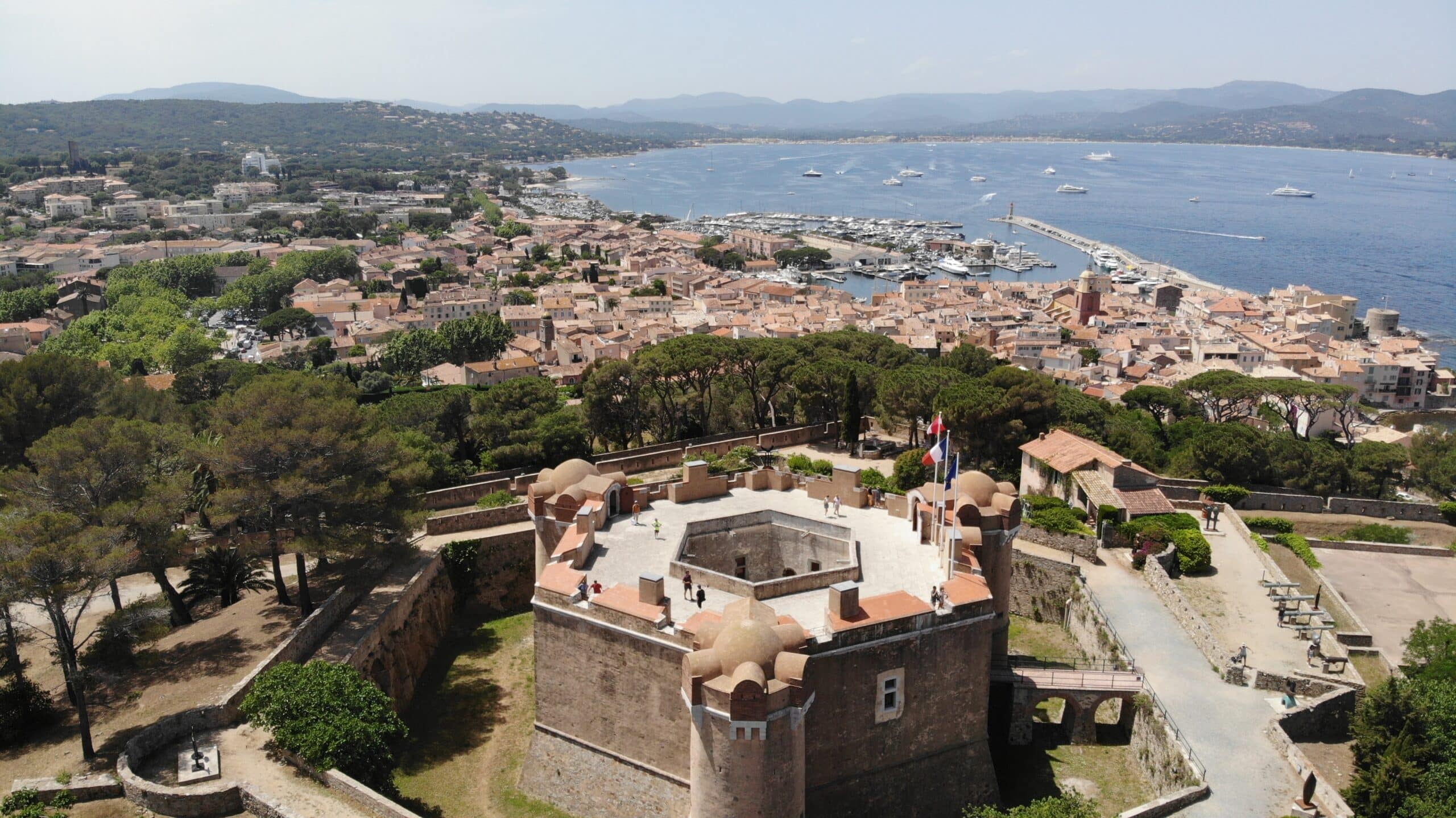 Luftaufnahme einer historischen Festung mit Türmchen über einer Küstenstadt mit einem Jachthafen, Hügeln und Booten auf dem Wasser im Hintergrund.