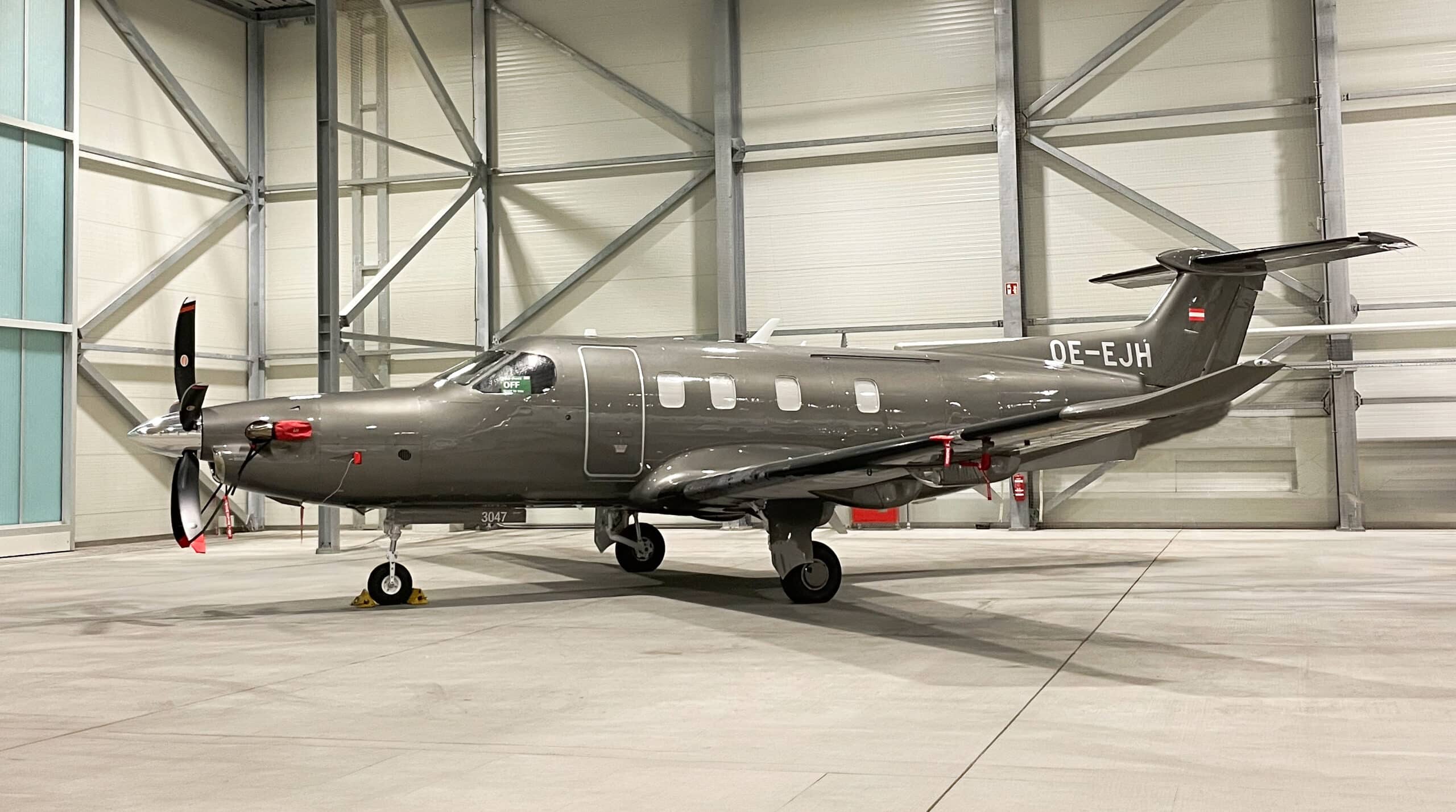 Ein graues einmotoriges Turboprop-Flugzeug mit dem Kennzeichen OE-EJH ist in einem geräumigen und gut beleuchteten Hangar geparkt.