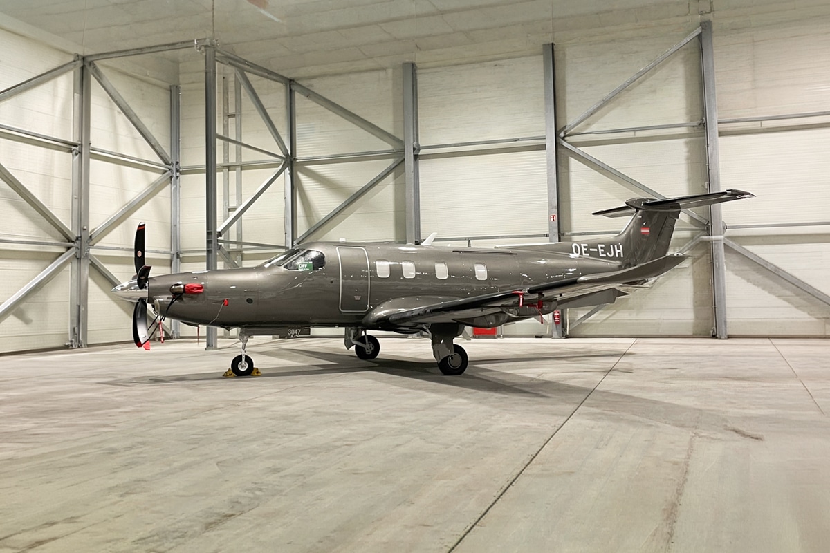 Ein graues einmotoriges Turboprop-Flugzeug mit dem Kennzeichen OE-EJH ist in einem geräumigen, leeren Hangar geparkt.