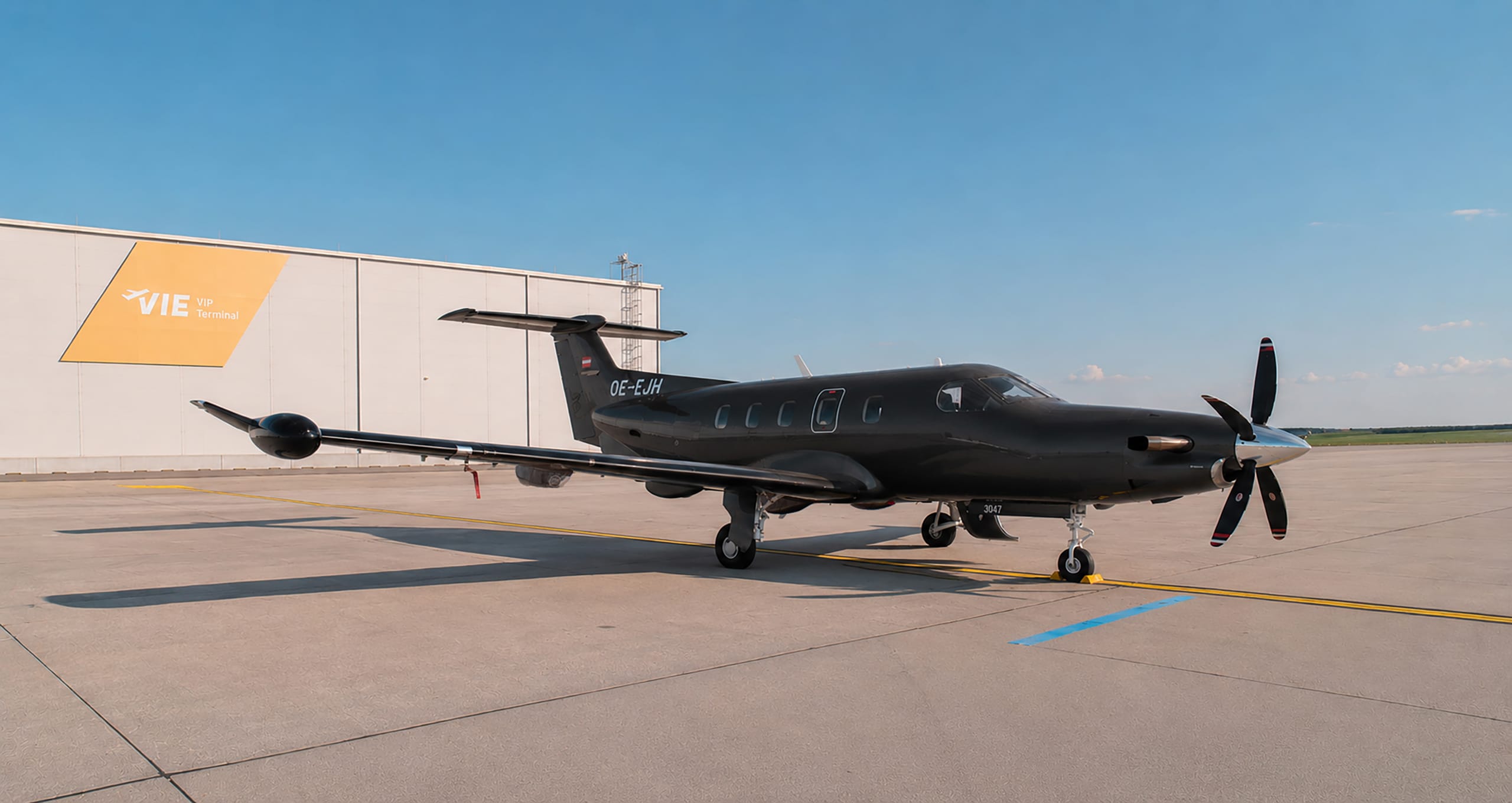 Ein schwarzes einmotoriges Turboprop-Flugzeug parkt auf der Rollbahn in der Nähe eines Gebäudes mit der Aufschrift "VIE VIP Terminal" unter einem klaren blauen Himmel.